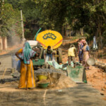 Bolpur, WB, India - November 30, 2018: Woman is carrying aggregates bowl on her head. Fellow laborers are working around concrete batch mixer. Everybody is dressed in usual clothes. In background rural road is under construction.